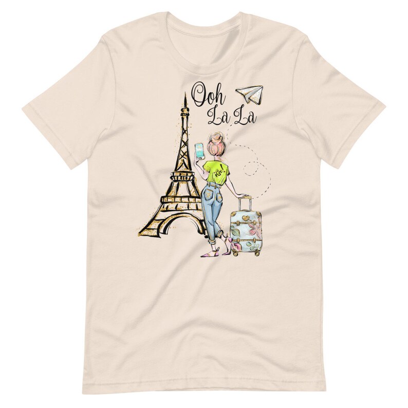 Op de afbeelding: Een beige t-shirt met een afbeelding van een vrouw die voor de Eiffeltoren staat met een koffer en een smartphone. De tekst "Ooh La La" staat boven de Eiffeltoren.