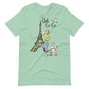 Op de afbeelding: Een groen T-shirt met een afbeelding van een vrouw die voor de Eiffeltoren staat met een koffer en een papieren vliegtuigje. De tekst "Ooh La La" staat boven de Eiffeltoren.