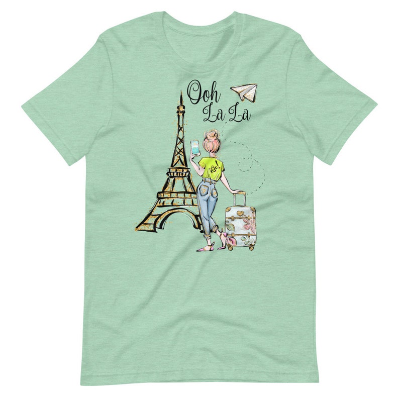 Op de afbeelding: Een groen T-shirt met een afbeelding van een vrouw die voor de Eiffeltoren staat met een koffer en een papieren vliegtuigje. De tekst "Ooh La La" staat boven de Eiffeltoren.