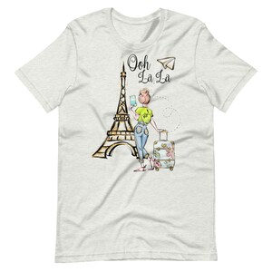 Op de afbeelding: Een wit T-shirt met een afbeelding van een vrouw die voor de Eiffeltoren staat met een koffer en een telefoon. De tekst "Ooh La La" staat boven de vrouw.
