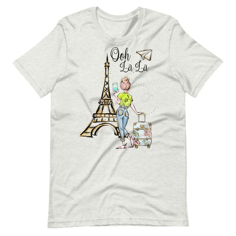 Op de afbeelding: Een wit T-shirt met een afbeelding van een vrouw die voor de Eiffeltoren staat met een koffer en een telefoon. De tekst "Ooh La La" staat boven de vrouw.