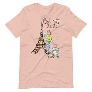Op de afbeelding: Een roze T-shirt met een afbeelding van een vrouw die voor de Eiffeltoren staat met een koffer en een papieren vliegtuig. De tekst "Ooh La La" staat boven de Eiffeltoren.