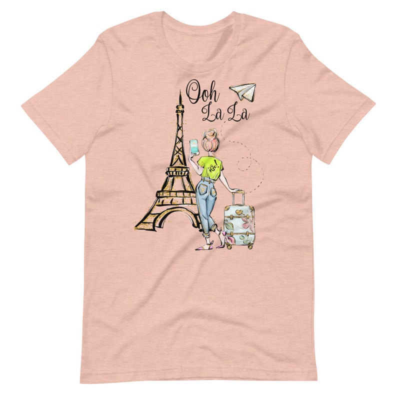 Op de afbeelding: Een roze T-shirt met een afbeelding van een vrouw die voor de Eiffeltoren staat met een koffer en een papieren vliegtuig. De tekst "Ooh La La" staat boven de Eiffeltoren.