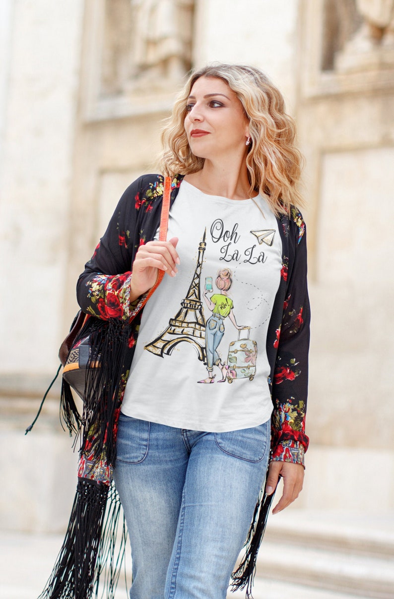 Op de afbeelding: Wit T-shirt met een afbeelding van de Eiffeltoren en een vrouw die een telefoon vasthoudt. De tekst "Ooh La La" is op het shirt gedrukt met een papieren vliegtuig.