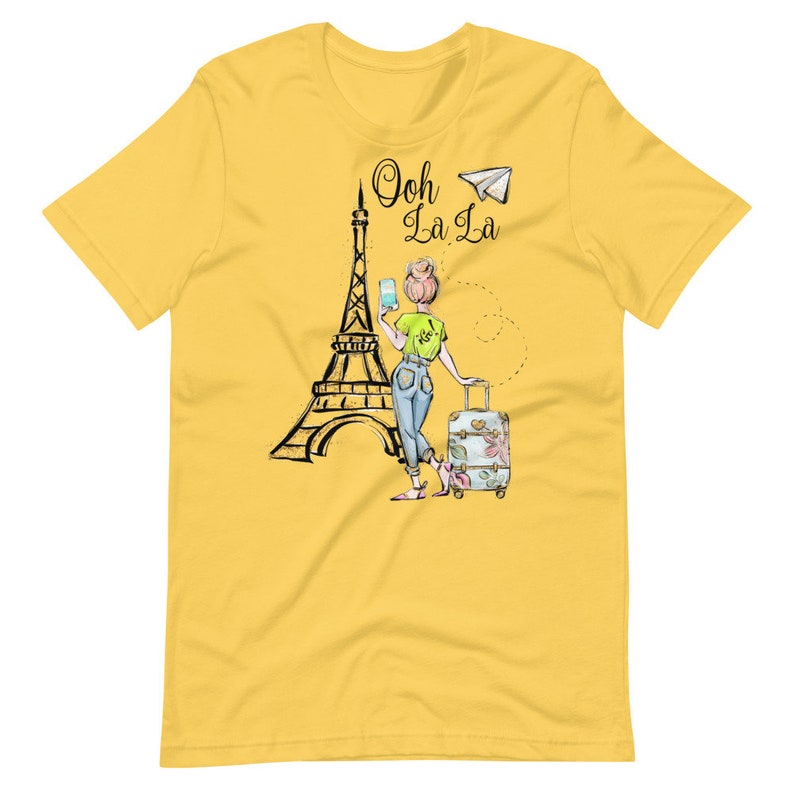 Op de afbeelding: Geel T-shirt met een afbeelding van een vrouw die voor de Eiffeltoren staat met een koffer en een papieren vliegtuigje. De tekst "Ooh La La" staat boven de Eiffeltoren.