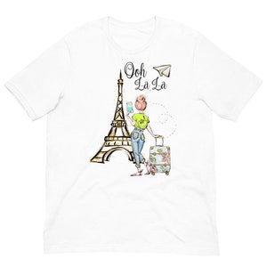 Op de afbeelding: Wit T-shirt met een afbeelding van een vrouw die voor de Eiffeltoren staat met een koffer en een telefoon. De tekst "Ooh La La" staat boven de afbeelding.