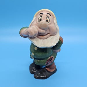 May include: A ceramic figurine of the character Sneezy from Snow White. The figurine is wearing a green coat, brown pants, and black shoes. The figure has a white beard and a yellow hat. The figurine is standing on a blue surface.