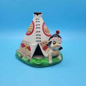 May include: A ceramic figurine featuring a white teepee with red and yellow accents, and a small Native American figure. The figurine is set on a green base and has a blue background.