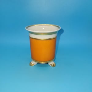 May include: A small porcelain vessel featuring a vibrant orange body with a white upper rim. The piece is accented with thin gold bands around the top and middle sections and is supported by three small, gold-trimmed feet at the base.