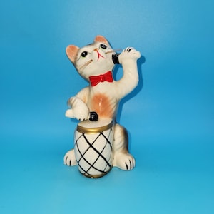 Puede incluir: Una figurita de cerámica de un gato tocando la batería. El gato lleva una pajarita roja y tiene un micrófono en su pata derecha. El tambor es negro y blanco con un borde dorado.