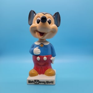 May include: A ceramic figurine of Mickey Mouse wearing a blue shirt and red pants. The figurine is standing on a base that says "Walt Disney World".