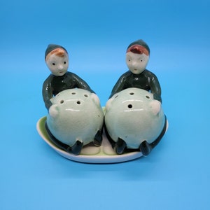 May include: A set of two ceramic salt and pepper shakers shaped like elves holding green pea pods. The elves have brown hair, green hats, and white faces.