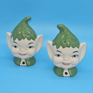 May include: A pair of vintage ceramic salt and pepper shakers. Each shaker is shaped like a smiling elf with a green hat and collar, white face, and large ears. The shakers have black eyes and small pink lips. The background is a solid blue.