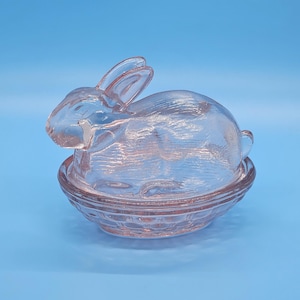 May include: A pink glass bunny-shaped trinket dish with a textured basket-weave base. The bunny has upright ears and is sitting atop the dish. The dish is a light pink color and is set against a blue background.