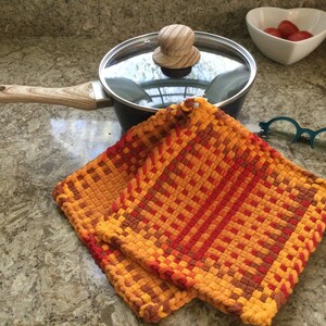 May include: Two orange, red, and brown woven pot holders on a granite countertop next to a black pot with a glass lid and a wooden knob.