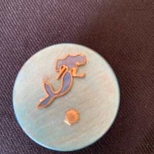 May include: A round wooden box with a blue and white painted surface. The box features a gold-coloured mermaid with a blue tail and a gold-coloured seashell.