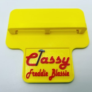 May include: A yellow plastic phone stand with a raised platform and a small sign that says "Classy Freddie Blassie".