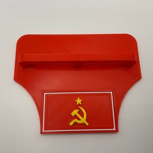 May include: Red 3D printed wall shelf with a white border and a red and yellow hammer and sickle emblem inside. The shelf has a small ledge on the top.