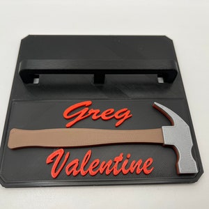 May include: A black wall mount with a silver hammer and the name "Greg Valentine" in red lettering.