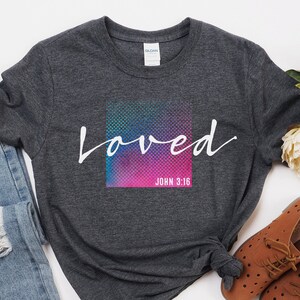 Pu&ograve; includere: Una maglietta grigia scura con una grafica bianca che dice "loved" con uno sfondo colorato e il testo "John 3:16" sotto. La maglietta &egrave; abbinata a jeans blu e scarpe marroni.