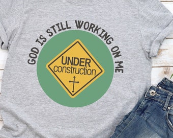 May include: A gray t-shirt with the text "God is still working on me" in a circle around a yellow construction sign with the text "Under construction" and a cross.