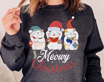 Peut inclure: Un sweat-shirt gris foncé avec un motif festif représentant trois chats de dessin animé portant des bonnets de Père Noël et tenant des cadeaux. Le texte "Meowy Christmas" est imprimé sous les chats.