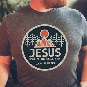 May include: A heather grey t-shirt with a black and orange graphic of a mountain range, a sun, and trees. The graphic is in a circle with the text "JESUS WENT TO THE WILDERNESS LUKE 5:16".