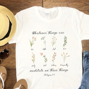K&ouml;nnte beinhalten: Wei&szlig;es T-Shirt mit einem floralen Design und dem Text "Whatever things are... meditate on these things - Philippians 4:8". Das Shirt liegt auf einer Holzoberfl&auml;che mit einem Strohhut, einer Sonnenbrille, Flip-Flops und einer Topfpflanze.