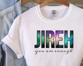 Può includere: Maglietta bianca con la parola "JIREH" stampata in lettere nere con un design di aurora boreale colorato dietro le lettere. Il testo "you are enough" è stampato in corsivo nero sotto la parola "JIREH".