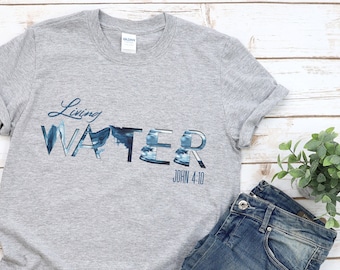 May include: Grey t-shirt with a blue and white graphic that says "Living Water" with the scripture reference "John 4:10" underneath. The shirt is laid out on a white wooden surface with a pair of orange sandals and blue jeans.
