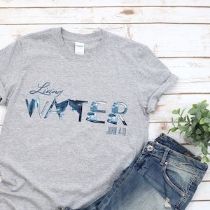 May include: Gray t-shirt with a blue and white graphic that says "Living Water" with the scripture reference "John 4:10" underneath. The shirt is laid out on a white wooden surface with a pair of orange sandals and blue jeans.