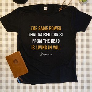 May include: Black t-shirt with a white graphic that reads "THE SAME POWER THAT RAISED CHRIST FROM THE DEAD IS LIVING IN YOU. Romans 8:11"