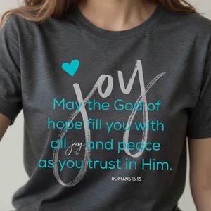 Op de afbeelding: Donkergrijs t-shirt met het woord "joy" in wit script en een turquoise hart. Het shirt bevat de tekst "Moge de God van de hoop u vervullen met alle vreugde en vrede, als u in Hem gelooft." uit Romeinen 15:13.