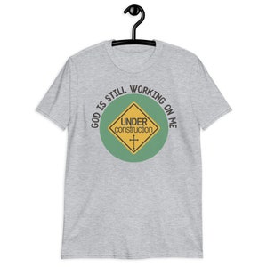 May include: A heather grey t-shirt with a green circle and a yellow construction sign that says "Under Construction" with a cross inside. The text around the circle says "God is still working on me".