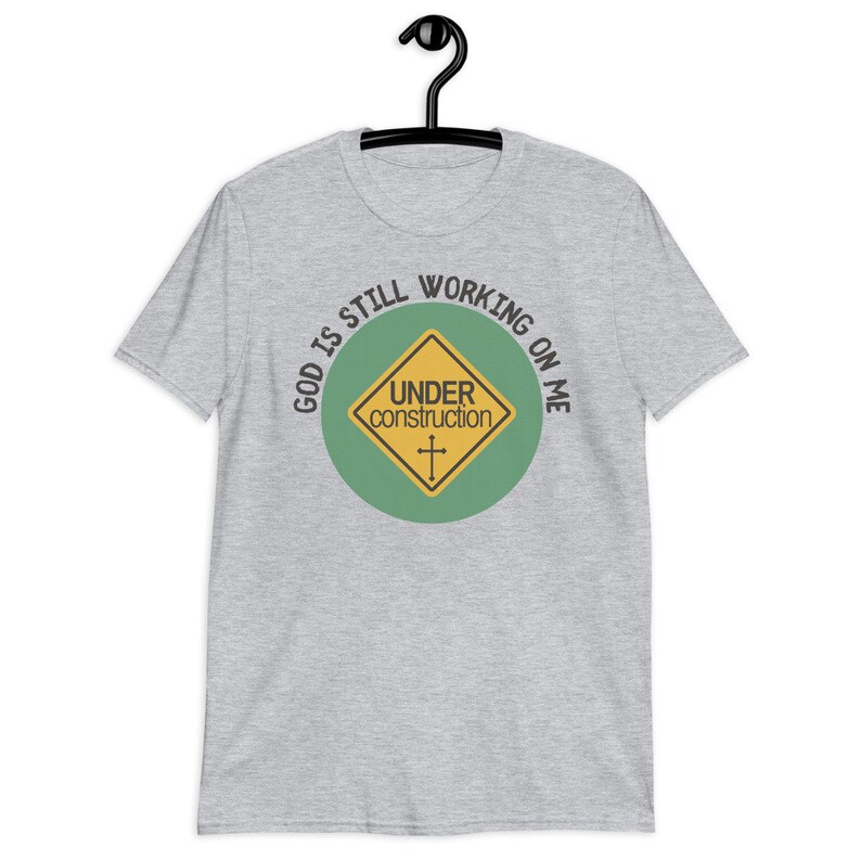 May include: A heather grey t-shirt with a green circle and a yellow construction sign that says "Under Construction" with a cross inside. The text around the circle says "God is still working on me".