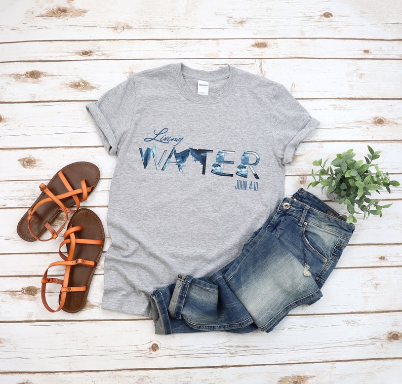 May include: Gray t-shirt with a blue and white graphic that says "Living Water" with the scripture reference "John 4:10" underneath. The shirt is laid out on a white wooden surface with a pair of orange sandals and blue jeans.