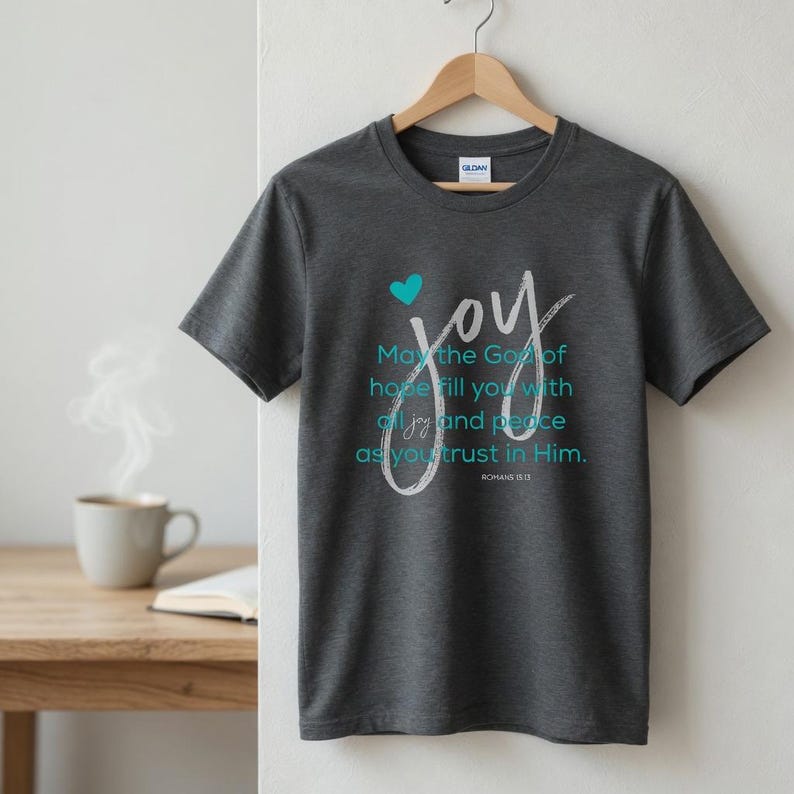 May include: Dark gray t-shirt with the word "joy" in white script and a teal heart. The shirt also features the text "May the God of hope fill you with all joy and peace as you trust in Him." ROMANS 15:13.