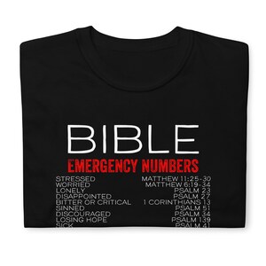 Puede incluir: Camiseta negra con texto blanco que dice "BIBLE EMERGENCY NUMBERS". El texto est&aacute; en negrita, sin serifas. Debajo del texto hay una lista de palabras en rojo, como "estresado", "preocupado", "solo" y "decepcionado", emparejadas con vers&iacute;culos b&iacute;blicos correspondientes en negro.