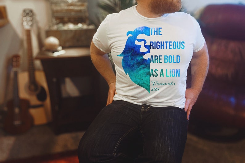 Pode incluir: Uma camiseta branca com um design de aquarela azul e verde de uma juba de le&atilde;o. O texto "The Righteous Are Bold As A Lion Proverbs 28:1" est&aacute; impresso na camiseta.