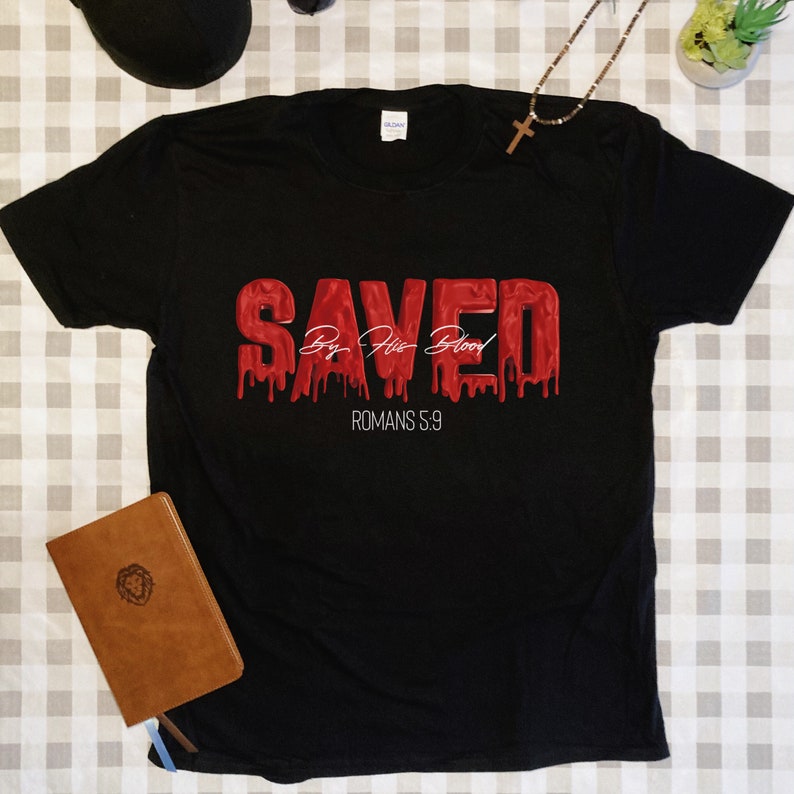 May include: Black t-shirt with a red graphic that says "SAVED By His Blood" and "ROMANS 5:9" in white text.