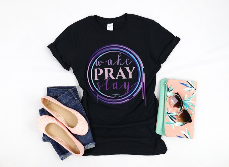 May include: Black t-shirt with a circle graphic that says "wake pray slay" in pink and purple lettering. The circle is outlined in blue, purple, and pink. The shirt is laid flat on a white surface with a pair of pink ballet flats and a floral clutch.