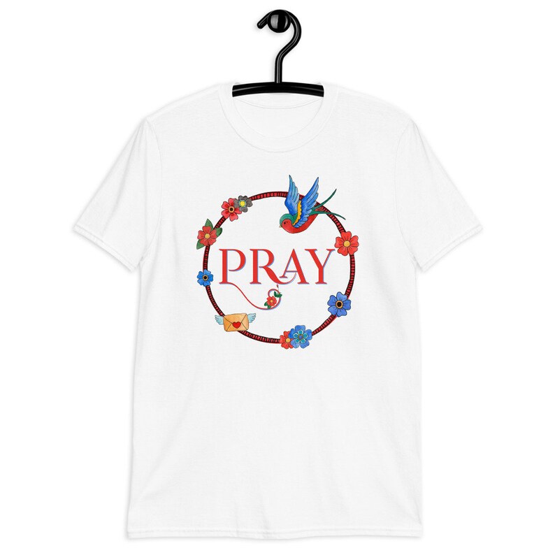 May include: White t-shirt with a red circle design featuring the word "PRAY" in red lettering, a blue and green bird, and colourful flowers.