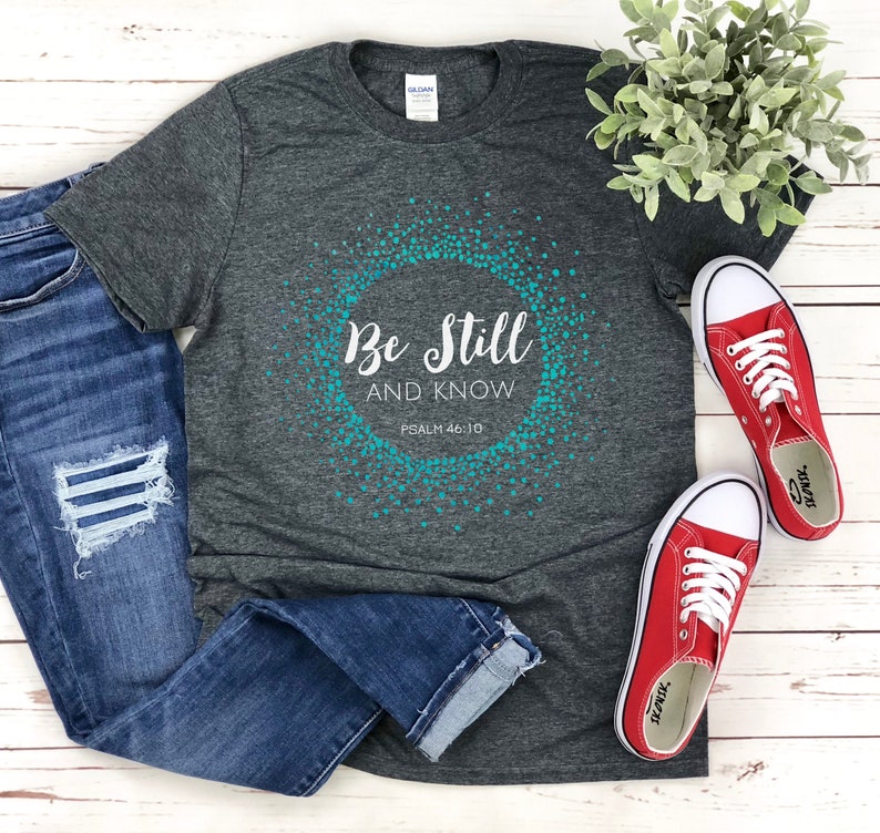 May include: A dark gray t-shirt with a teal blue circle design that says "Be Still and Know" with the text "Psalm 46:10" underneath. The shirt is laying on a white wooden surface with a pair of red converse shoes and blue jeans.