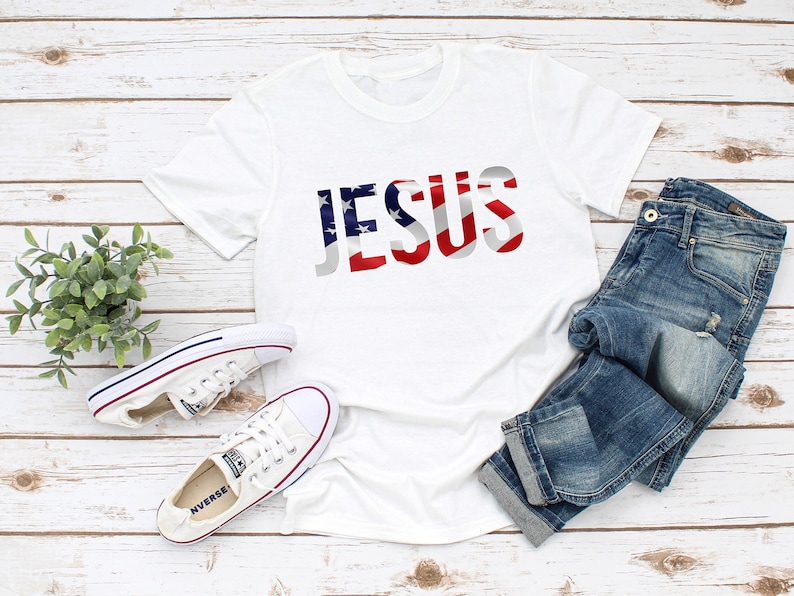 May include: White t-shirt with the word "JESUS" printed in red, white, and blue, resembling the American flag. The shirt is laid out on a white wooden surface with a pair of white sneakers and blue jeans.