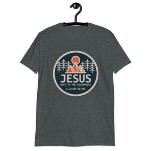 May include: A dark heather grey t-shirt with a circular graphic of a mountain range, a sun, and pine trees. The graphic has the text "JESUS WENT TO THE WILDERNESS LUKE 5:16".