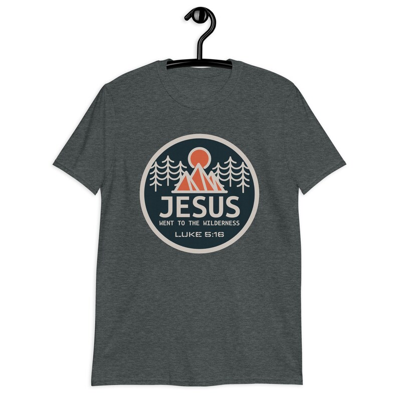 May include: A dark heather grey t-shirt with a circular graphic of a mountain range, a sun, and pine trees. The graphic has the text "JESUS WENT TO THE WILDERNESS LUKE 5:16".