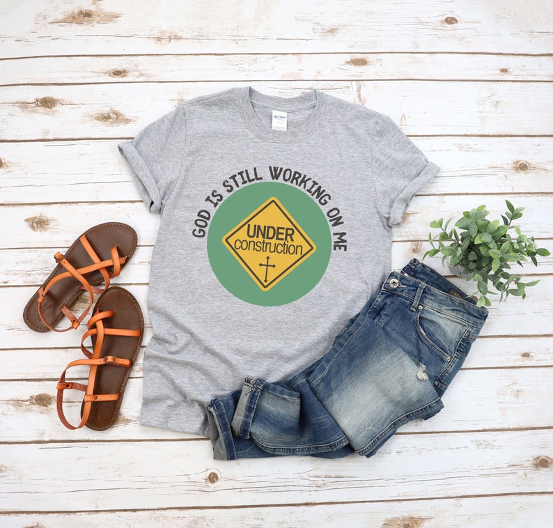 May include: A gray t-shirt with the text "God is still working on me" in a circle around a yellow construction sign with the text "Under construction" and a cross.