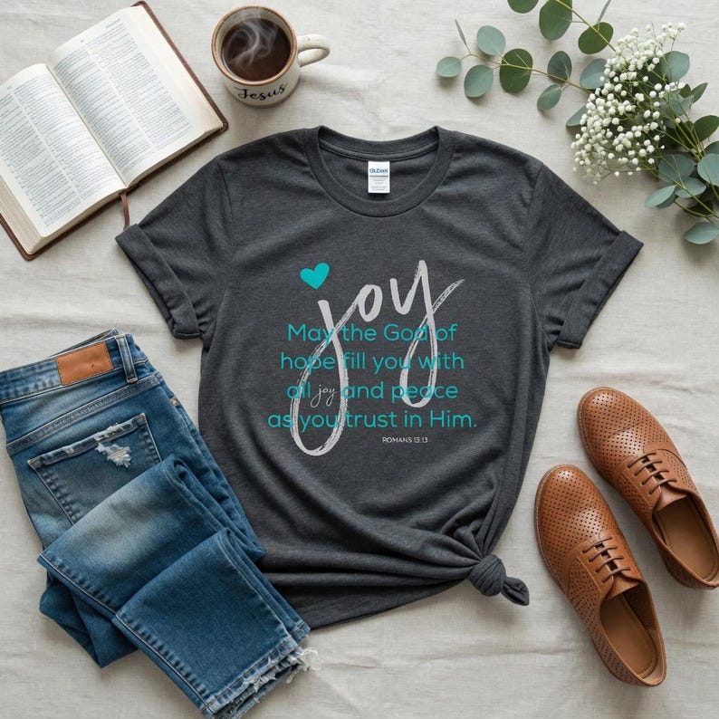 May include: Dark gray t-shirt with the word "joy" in white script and a teal heart. The shirt also features the text "May the God of hope fill you with all joy and peace as you trust in Him." Jeans and brown shoes are also visible.
