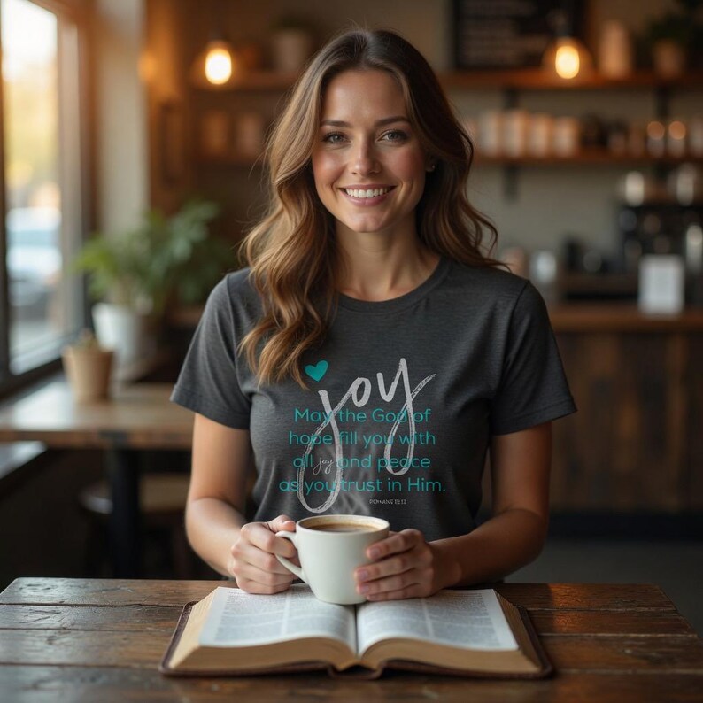 May include: A woman wearing a gray t-shirt with the word "joy" and a heart graphic. The shirt also features the text "May the God of hope fill you with all joy and peace as you trust in Him." She is holding a white mug and sitting at a table with an open book.