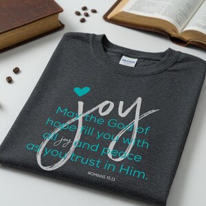 May include: Dark gray t-shirt with the word "joy" in white script and a teal heart. The shirt features the text "May the God of hope fill you with all joy and peace as you trust in Him." from Romans 15:13.